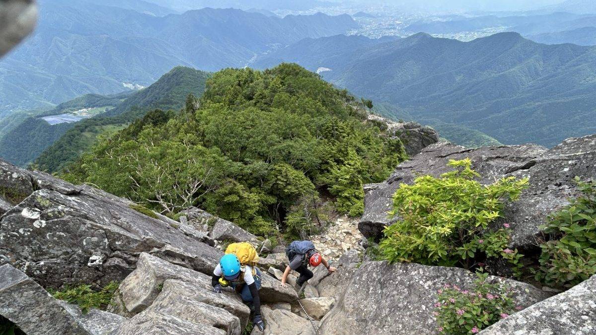 Group of climbers wearing helmets ascending a rocky mountain trail with a rope for safety, lush green hills in the background