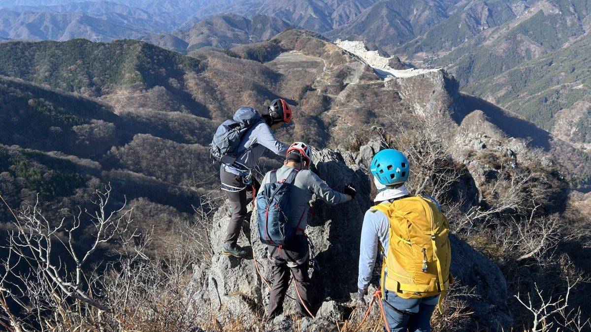 Group of climbers with helmets and backpacks standing on a rocky mountain ridge, overlooking distant mountain ranges.