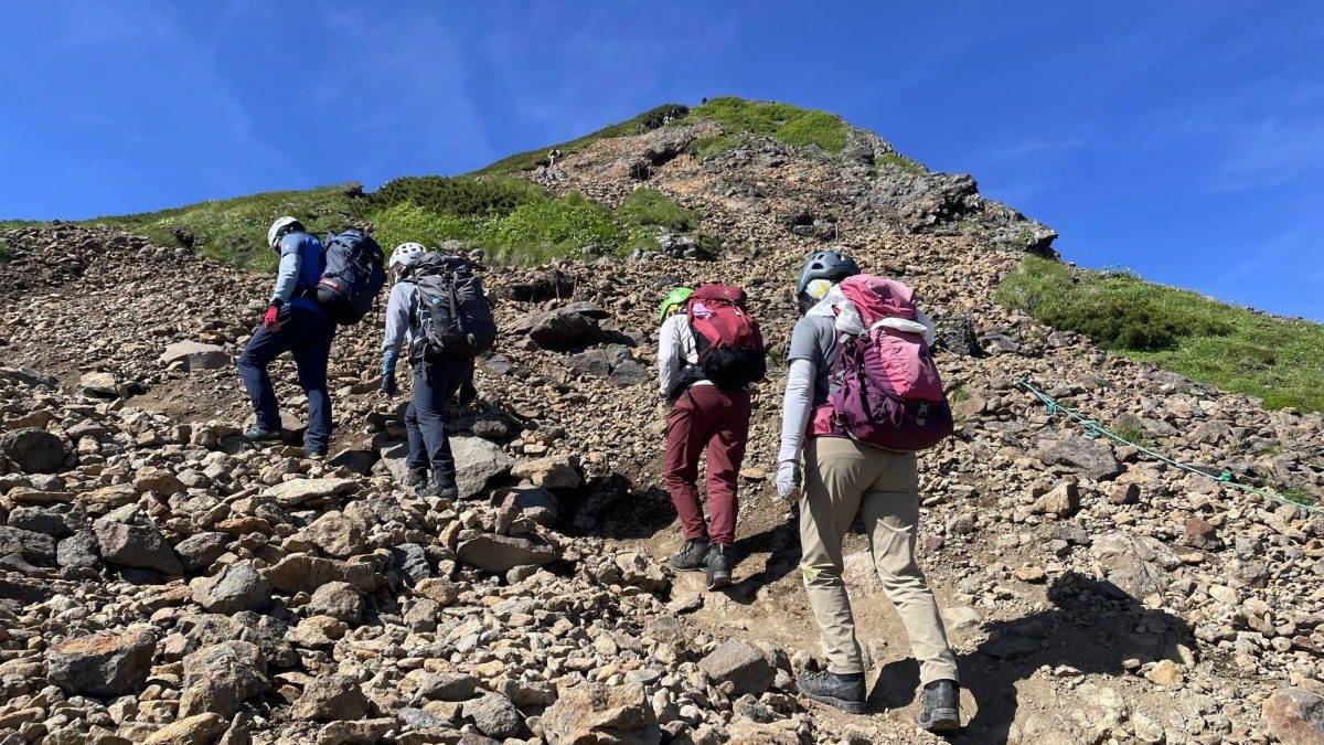Group of hikers wearing helmets and backpacks ascend a rocky mountain trail under a clear blue sky.