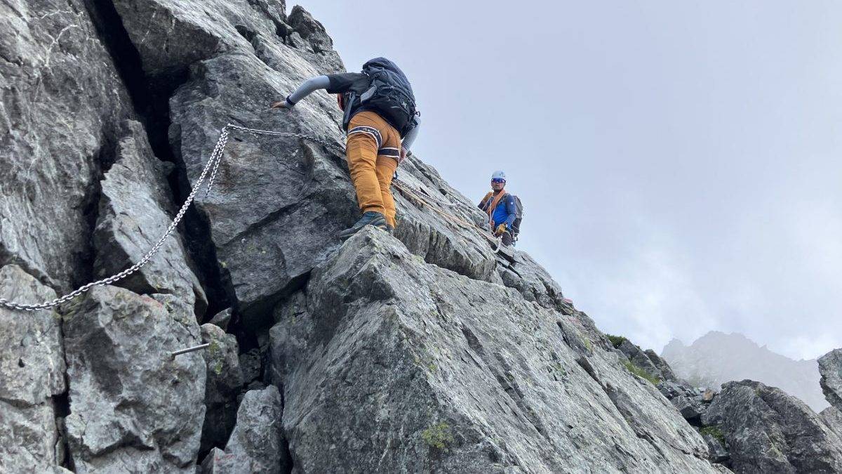 Mountaineers ascend steep rocky cliff using chain handholds, wearing helmets and backpacks.