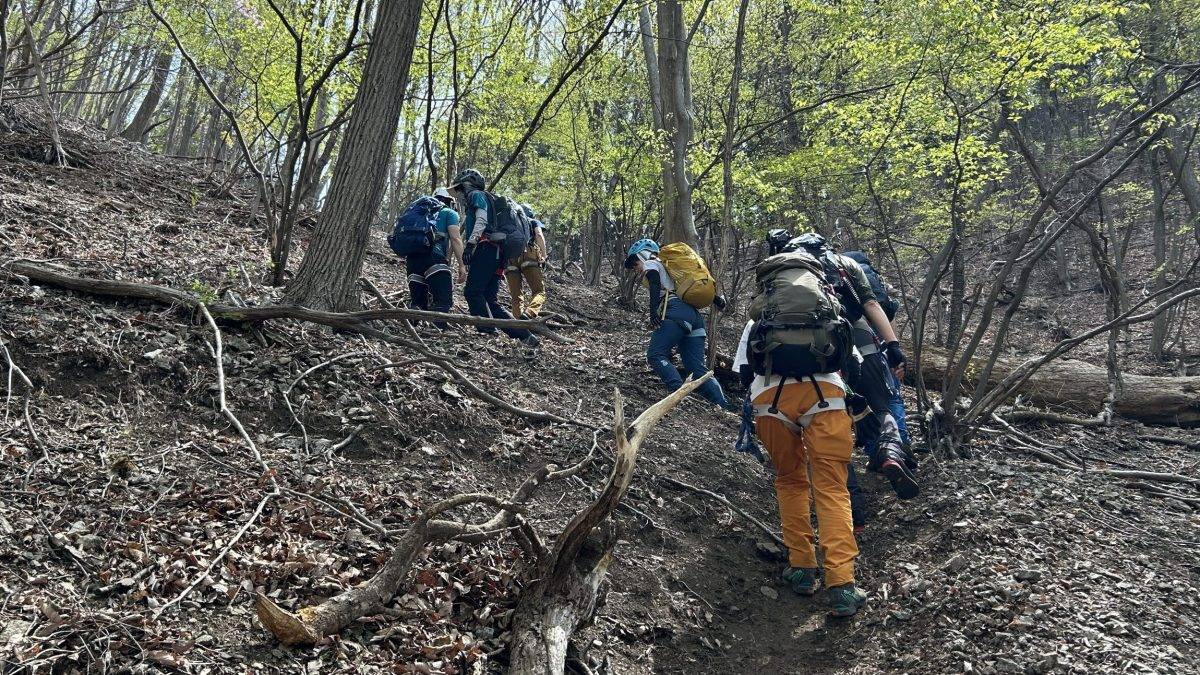 Group of hikers with backpacks and helmets ascending a rocky, leaf-strewn forest trail.