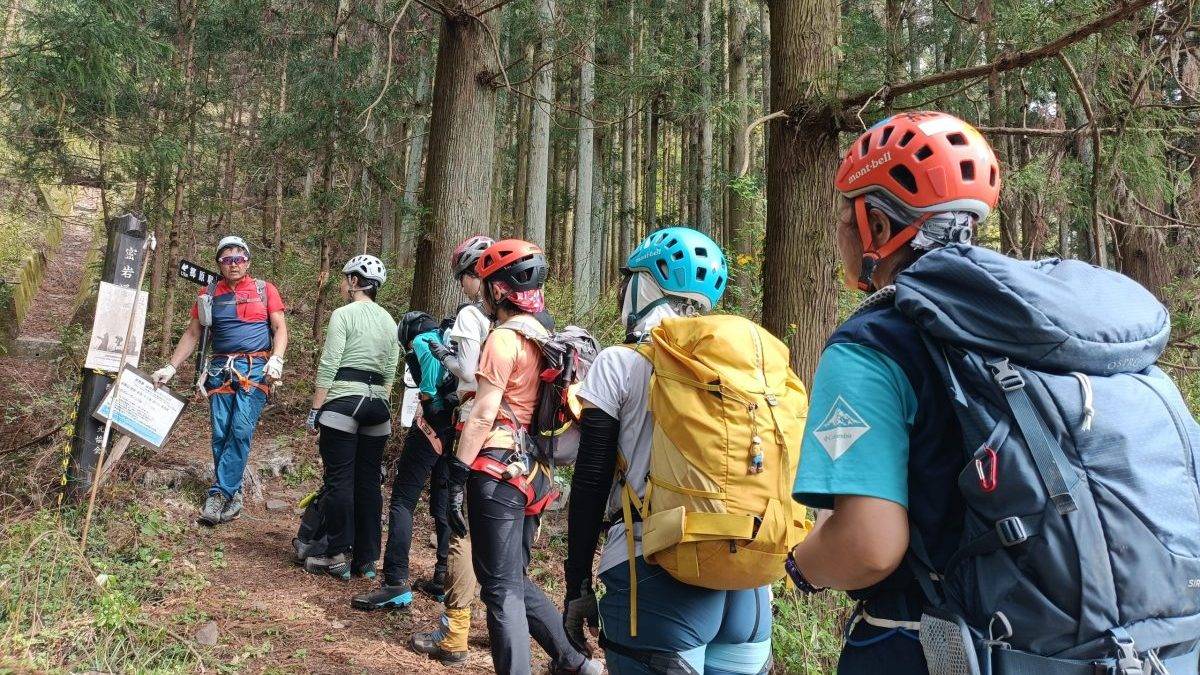 Group of hikers wearing helmets and backpacks stand on a dirt forest trail with signposts amid tall trees.