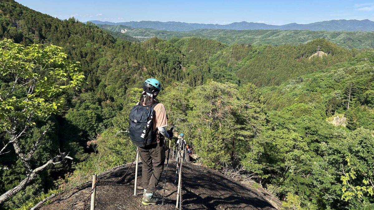 Hikers with helmets and backpacks walk along a rocky lava trail through forested hills and distant mountains under a clear blue sky.