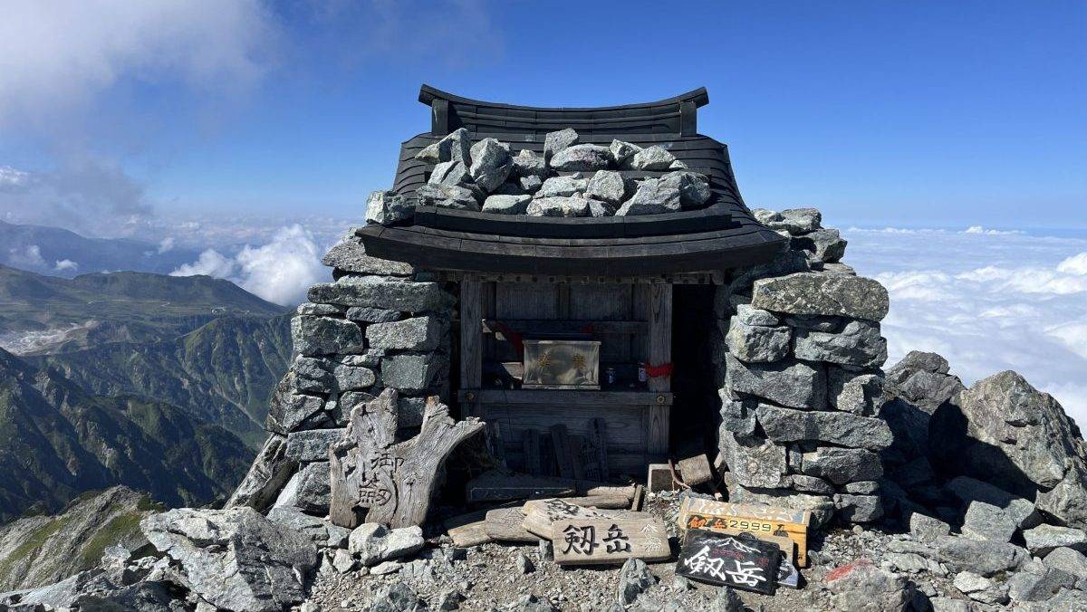 Stone mountain hut atop a rocky peak with a wooden roof, surrounded by piled rocks and signboards at the front. (informative)