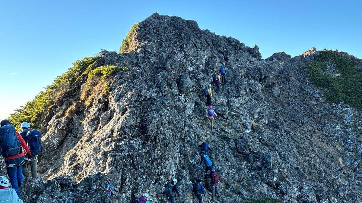 Hikers in helmets and backpacks ascending a jagged rocky mountain ridge against a clear blue sky.