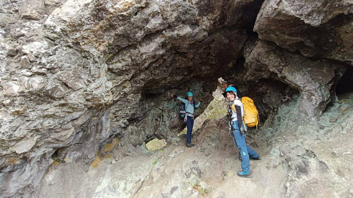 Group of hikers wearing helmets and backpacks standing inside a rocky cave or crevice with rough gray walls.