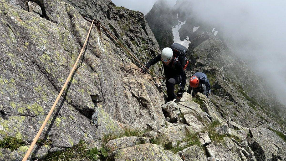 Climbers wearing helmets and backpacks ascend a jagged rock face on a misty mountain ridge, secured by ropes and chains.