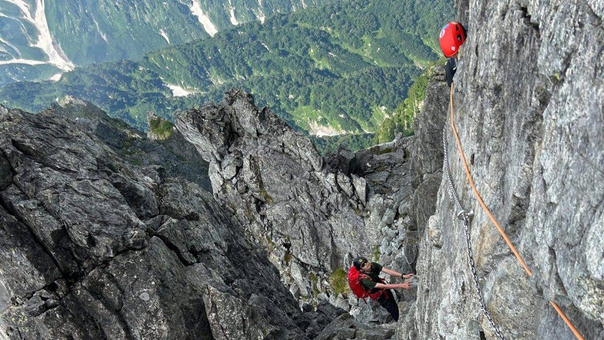 Rock climber wearing a red helmet ascending a jagged cliff with a steel chain anchor visible against a mountainous valley below