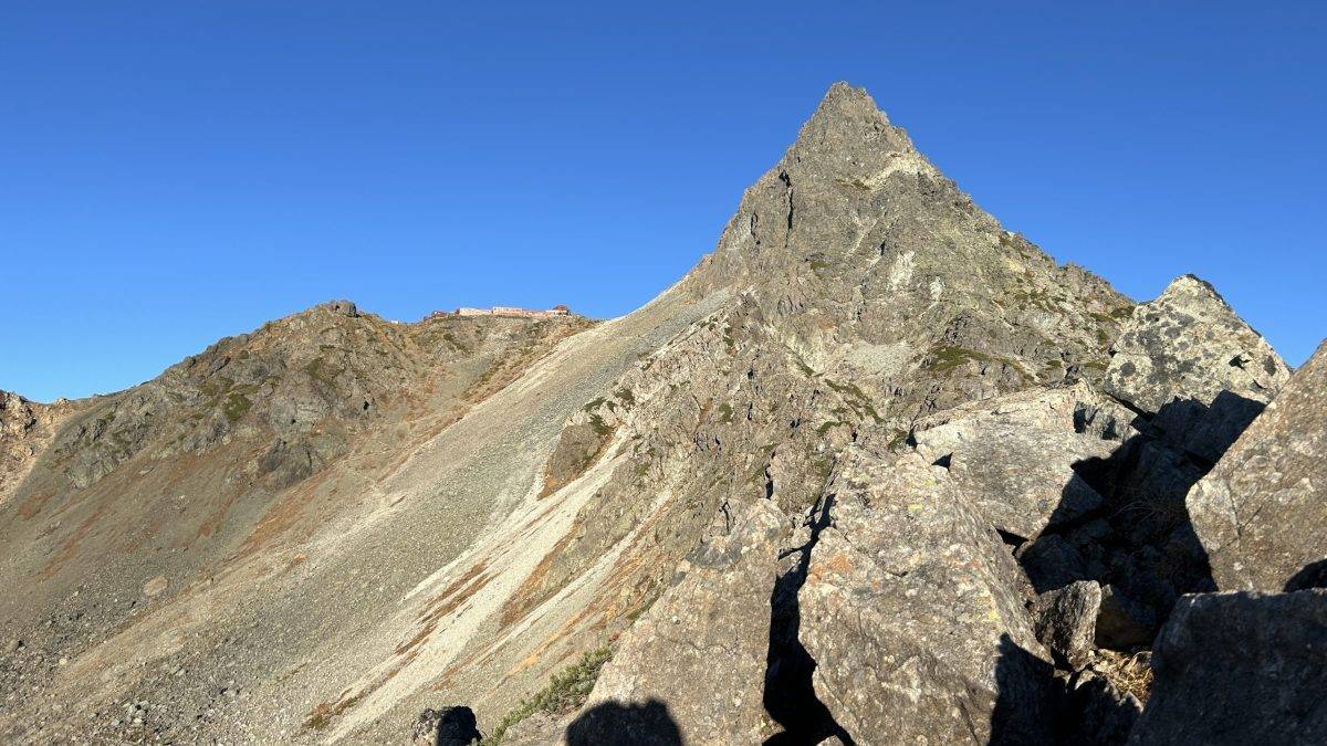 Rugged mountain ridge with a sharp peak under a clear blue sky; a small building sits on the left ridge.