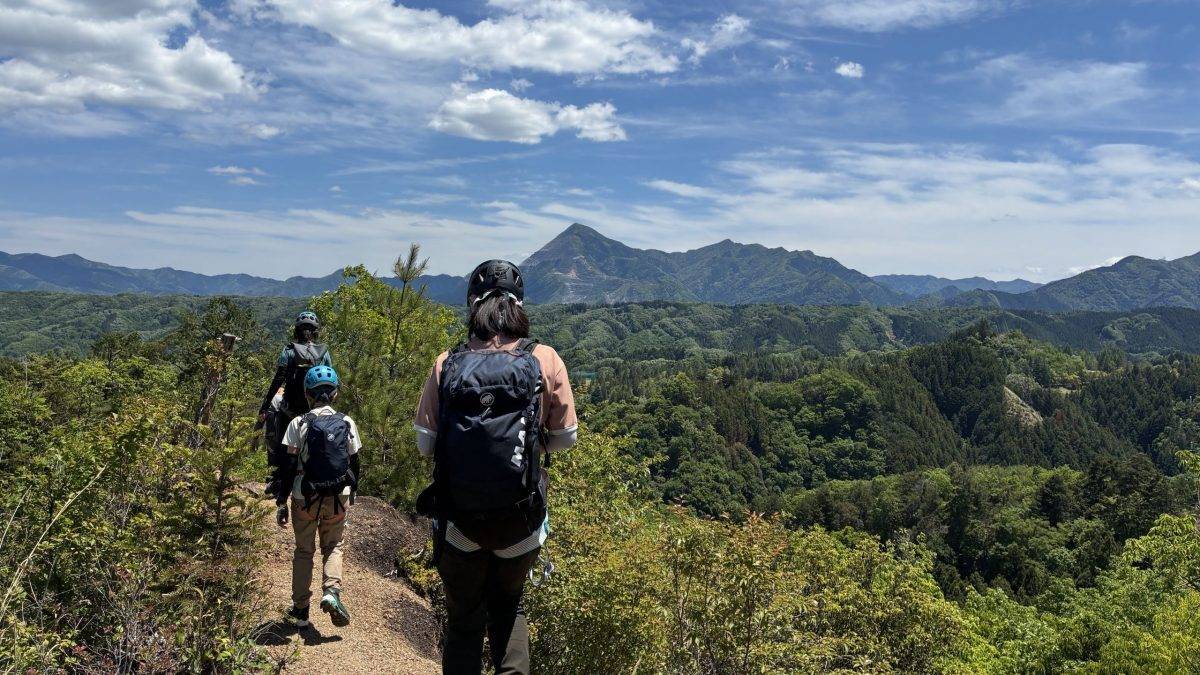Hikers wearing backpacks and helmets walk along a dirt trail with a mountain range and blue sky in the background.