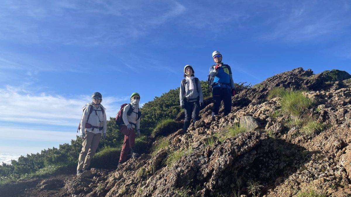 Group of hikers wearing helmets and backpacks standing on a rocky hillside under a clear blue sky.