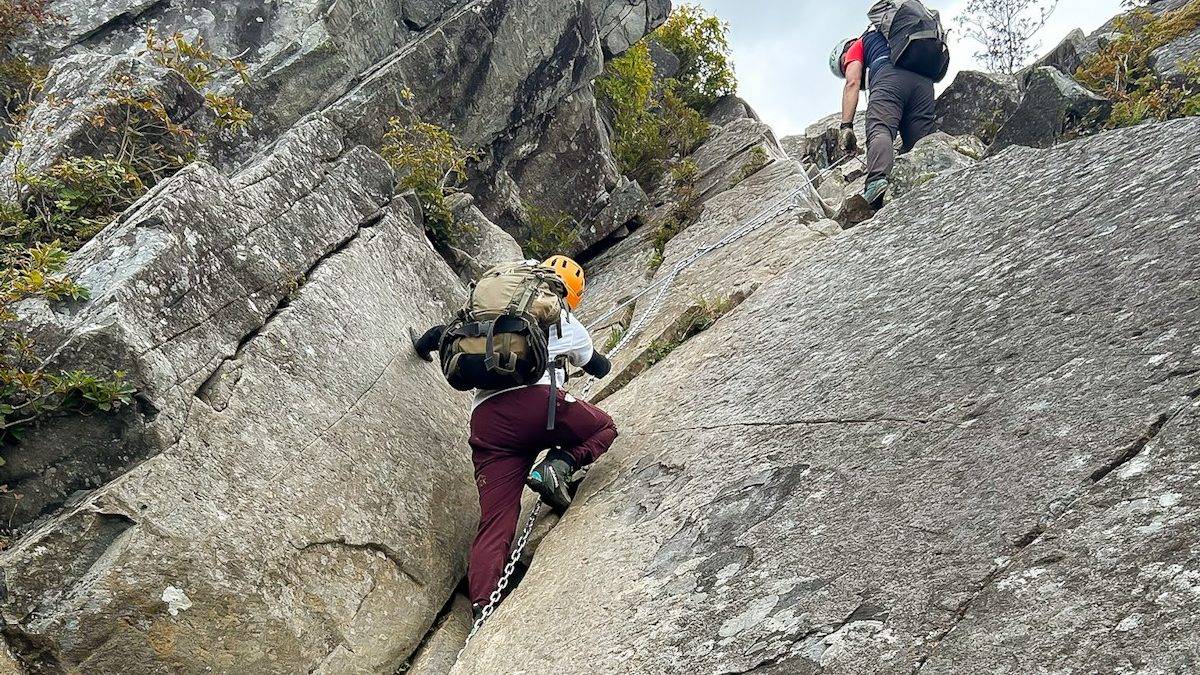 Two climbers ascending a steep rock face with a chain, wearing helmets and backpacks at a via ferrata-style route.