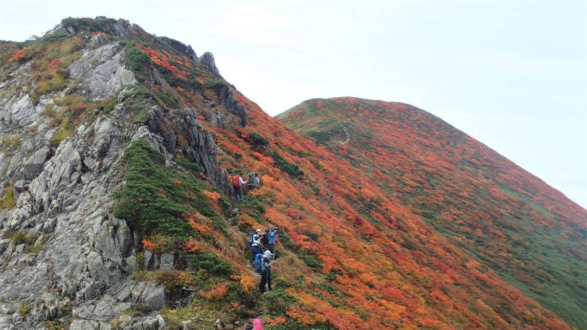 Hikers climbing a rocky mountain ridge covered in orange and red autumn foliage on a clear day.