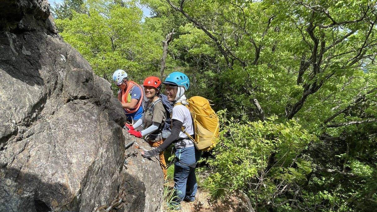Group of climbers wearing helmets and backpacks ascending a rocky cliff with dense green trees behind them.