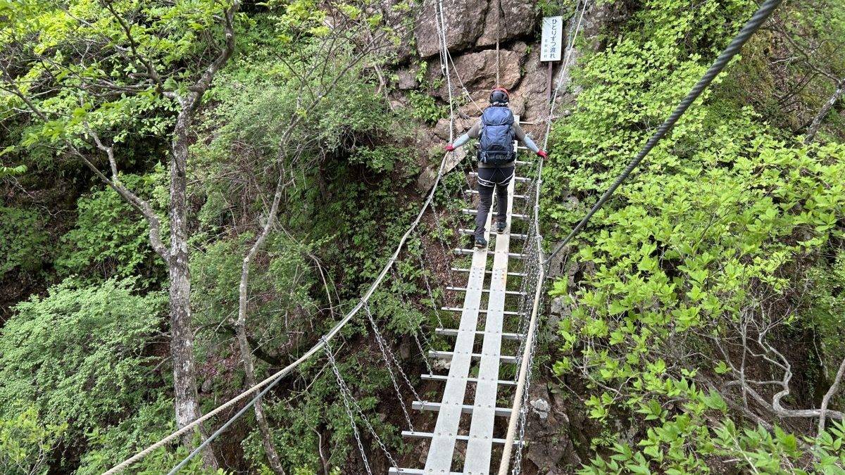 Hiker wearing a helmet and backpack crossing a narrow metal rope bridge through a green rocky ravine.