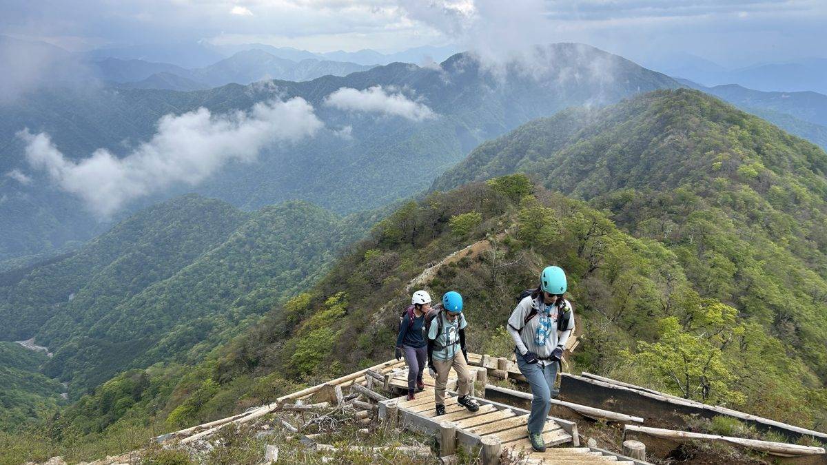Hikers wearing helmets walk a wooden boardwalk along a steep mountain ridge with misty, forested hills in the background.