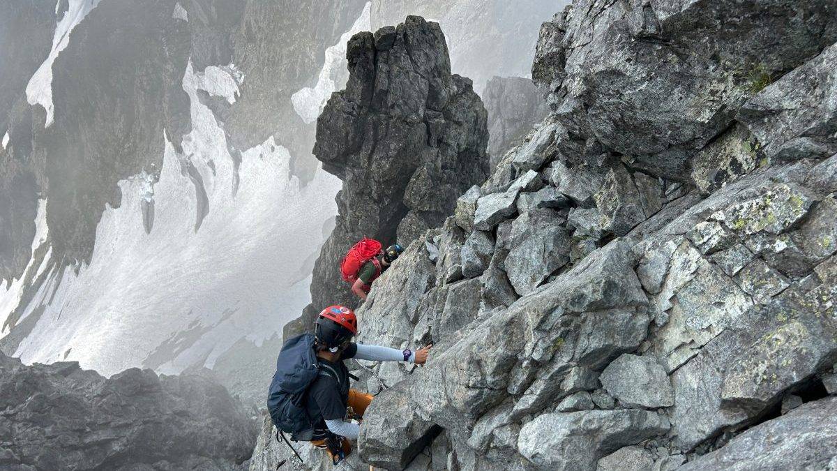 Mountaineers climbing a steep rock face with helmets and ropes in foggy, rocky terrain