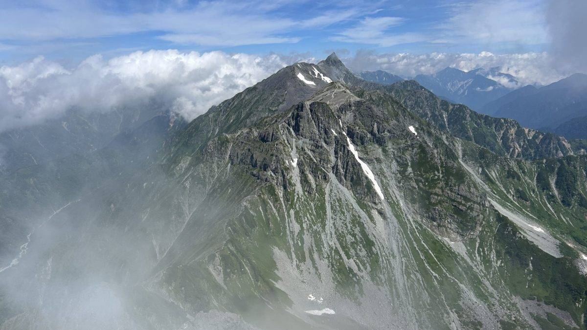 Jagged mountain peak rising above a cloudy, mist-filled valley under a blue sky with wispy clouds nearby mercurial ridges