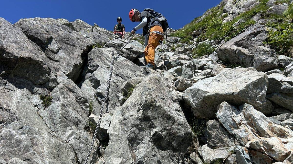 Two climbers in helmets navigate a jagged rocky slope using a metal chain, under a clear blue sky.
