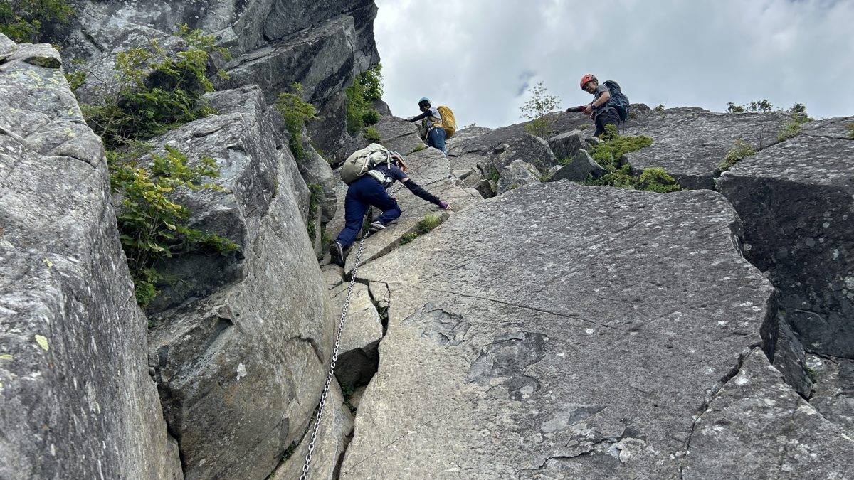 Mountaineers wearing helmets and backpacks climb a rugged rock face, using a safety chain for support.