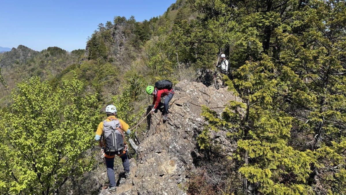 Hikers with helmets and backpacks traverse a rocky, cliffside trail on a forested mountainside under a clear blue sky.