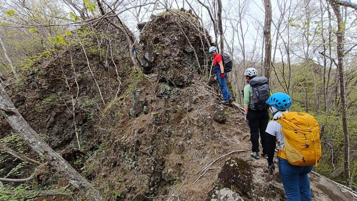 Hikers wearing helmets and backpacks navigate a rocky, narrow forest trail.