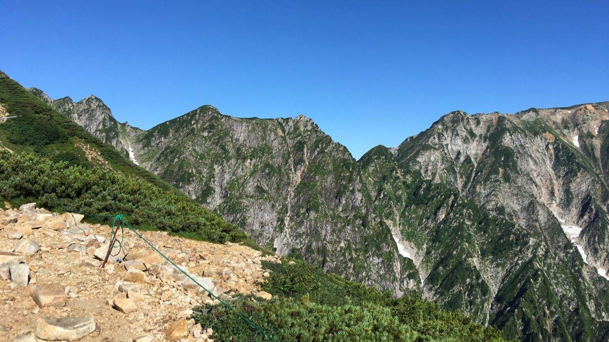 Rugged mountain ridge with rocky path in foreground, green shrubs, and a clear blue sky above