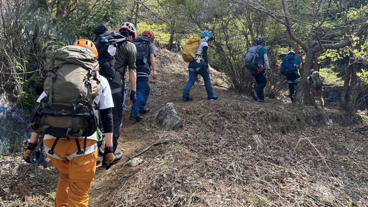 Group of hikers with backpacks and helmets walking uphill on a wooded trail