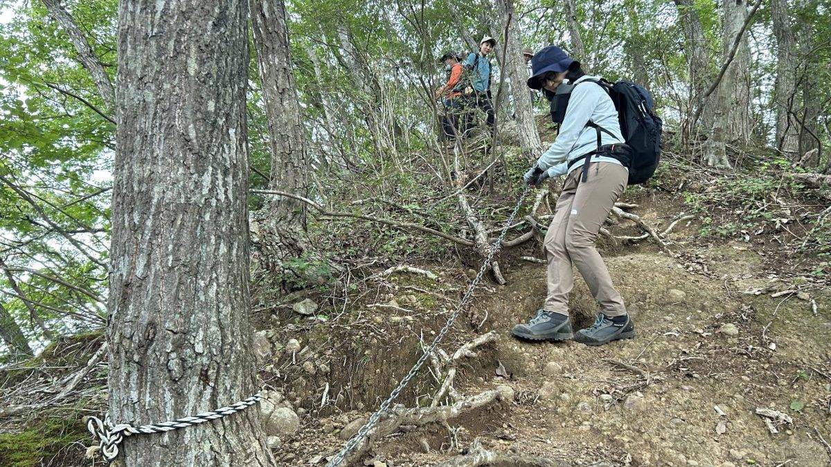 Hikers navigate a steep rocky trail with steel chains for handholds along a wooded slope, wearing backpacks and hiking gear.