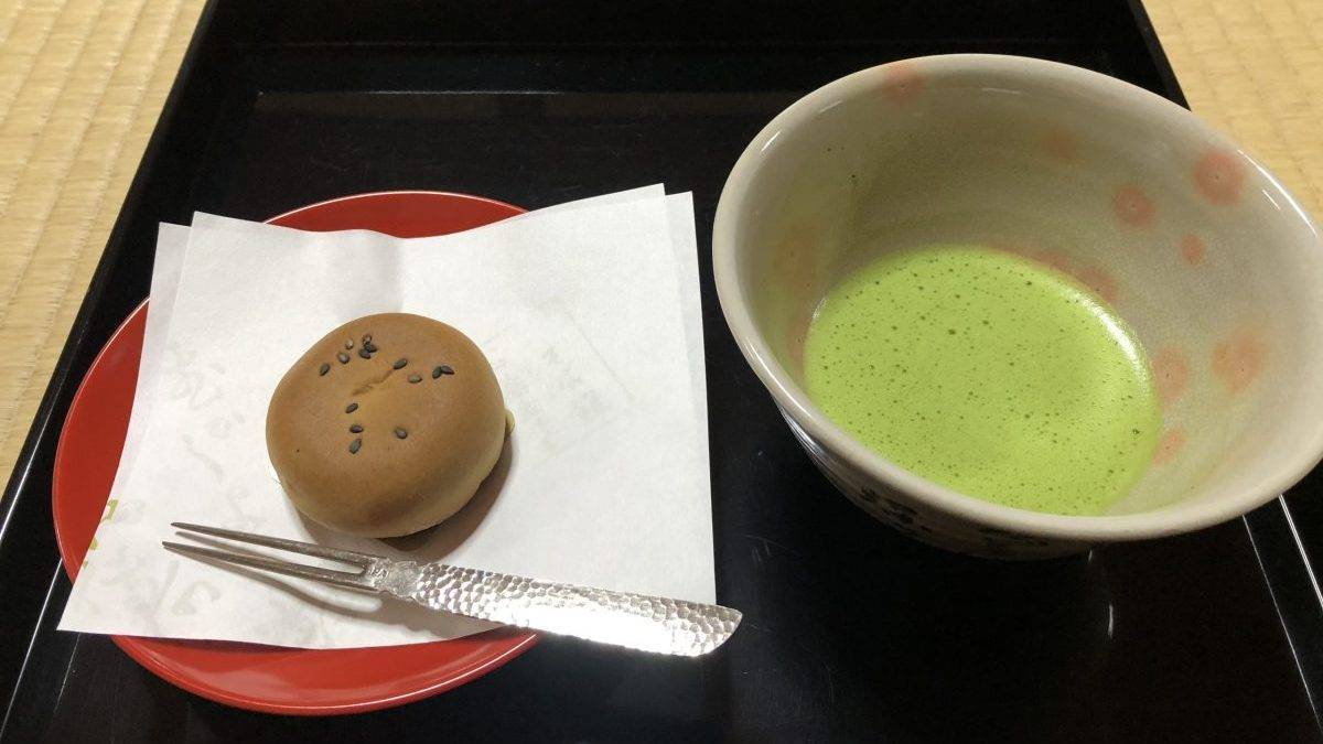 Matcha tea in a ceramic bowl on a black tray, with a sesame-topped bun on a napkin-covered red plate nearby.