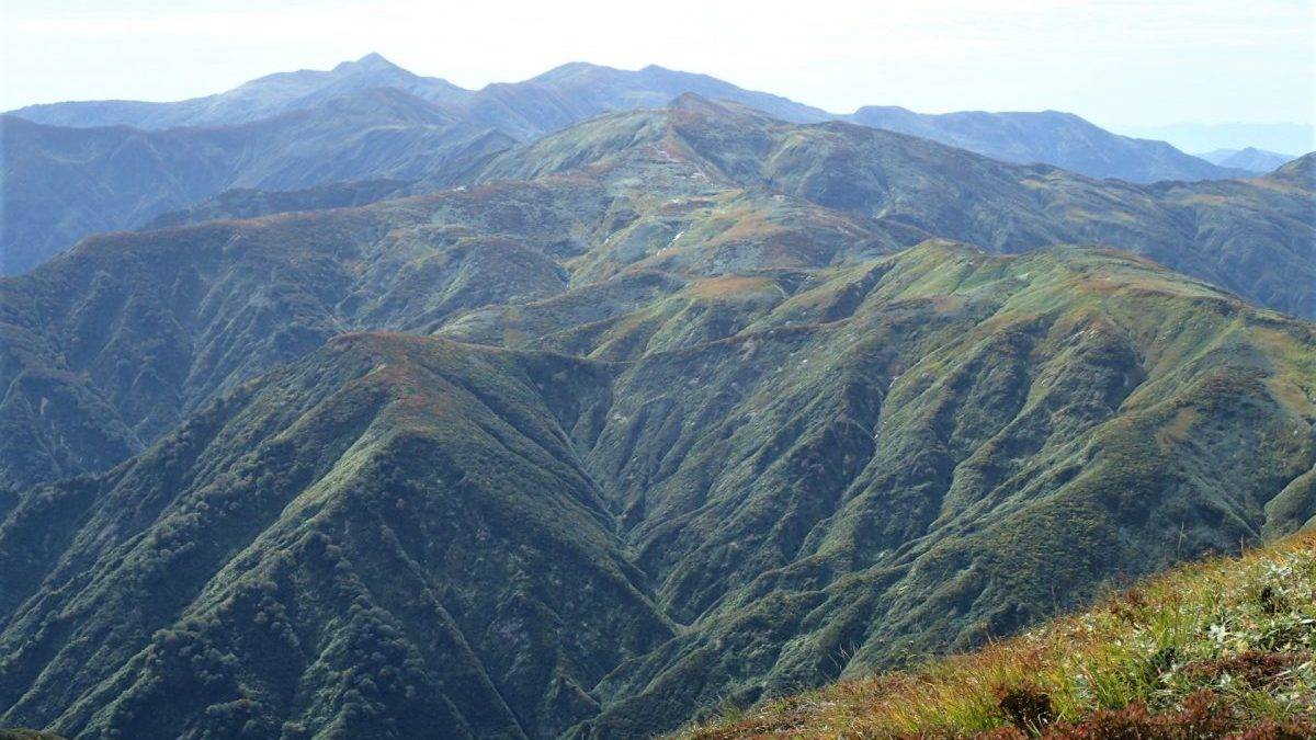 Wide view of rugged green mountains with ridges fading into the distance and a grassy slope in the foreground.