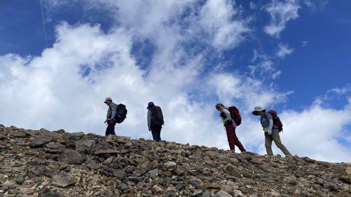 Group of hikers with backpacks ascending a rocky mountain slope under a blue sky with clouds.