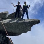 Two climbers in helmets and harnesses stand on a rock ledge, giving thumbs up amid a blue sky and clouds, with safety ropes nearby.