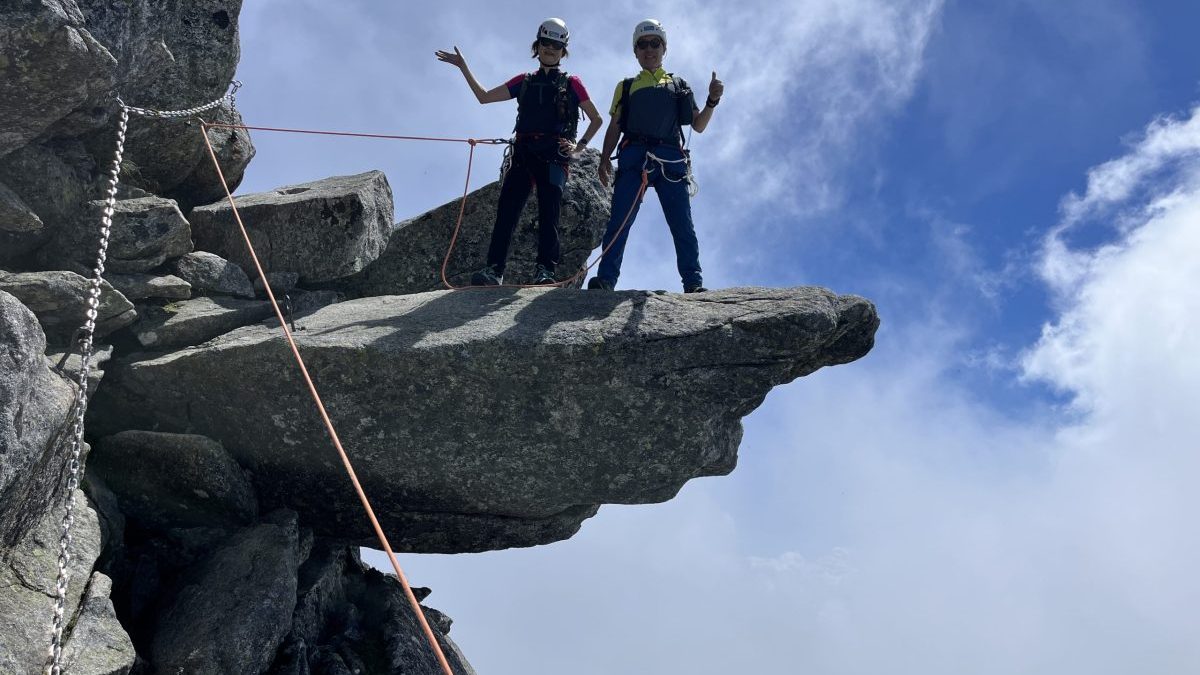 Two climbers in helmets and harnesses stand on a rock ledge, giving thumbs up amid a blue sky and clouds, with safety ropes nearby.