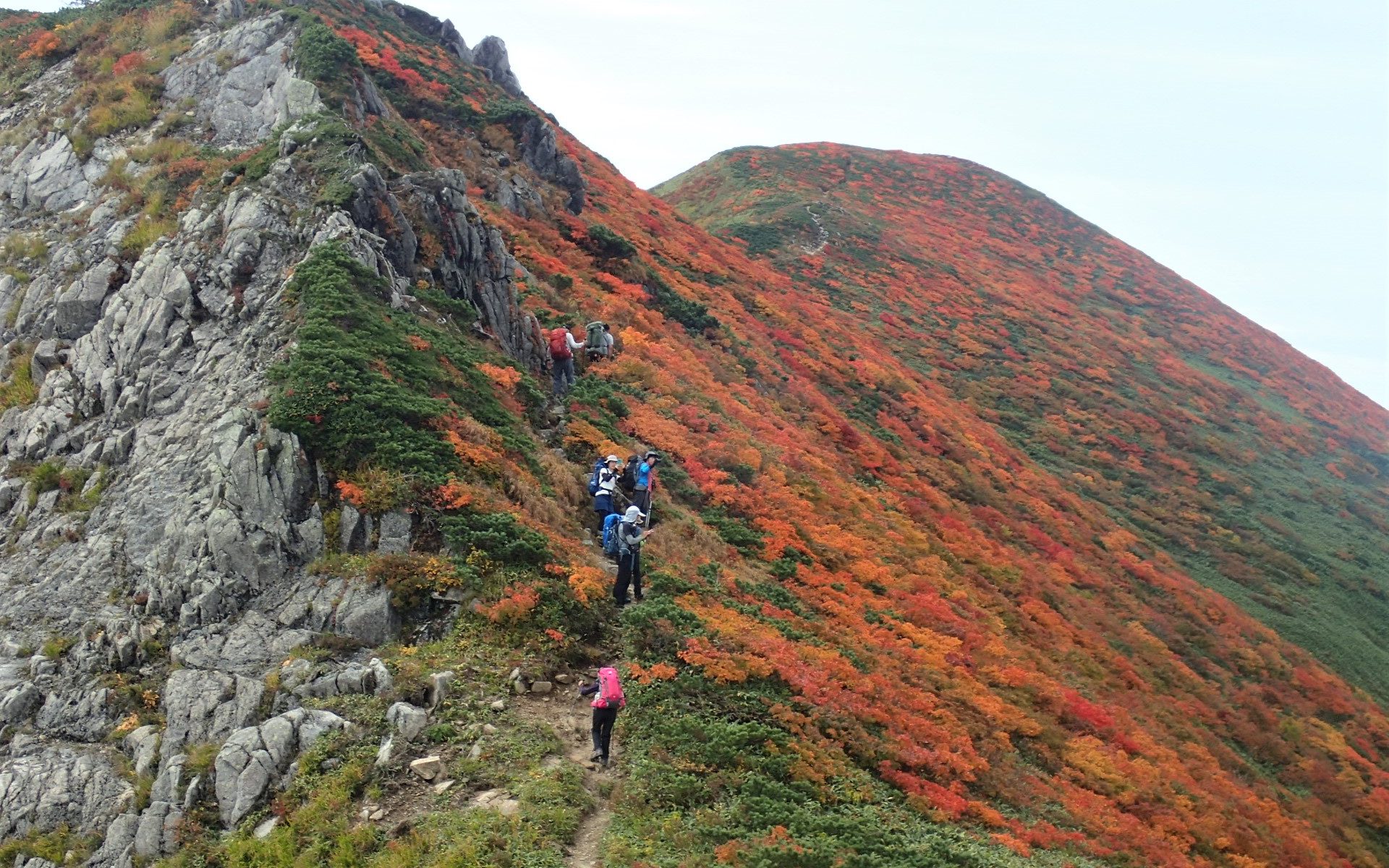 Hikers climbing a rocky mountain ridge covered in orange and red autumn foliage on a clear day.