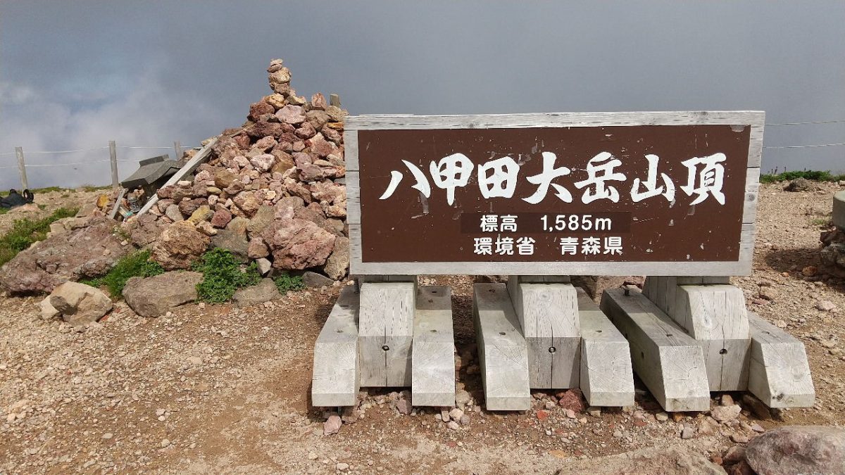 Sign at the summit of Mount Hakkoda with elevation 1,585 m, supported by concrete blocks and a stone cairn nearby.