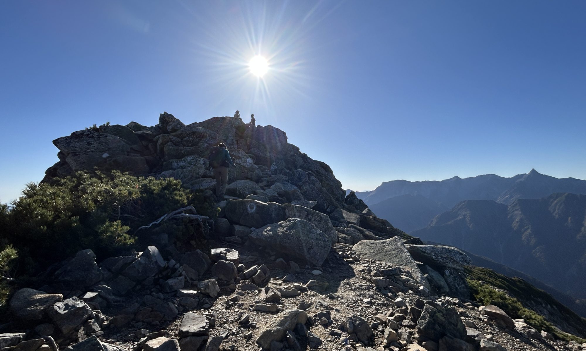 Hikers on a jagged rocky summit under a bright sun with a clear blue sky and distant mountain ranges behind them.