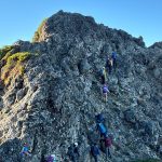 Hikers in helmets and backpacks ascending a jagged rocky mountain ridge against a clear blue sky.