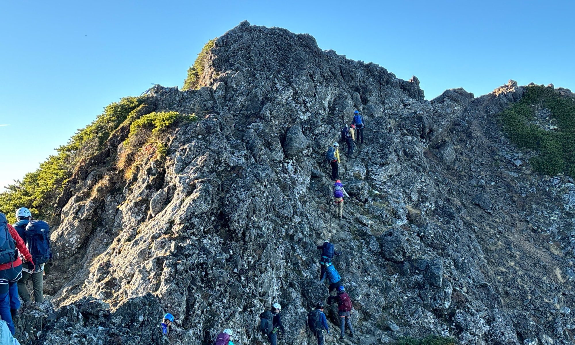 Hikers in helmets and backpacks ascending a jagged rocky mountain ridge against a clear blue sky.
