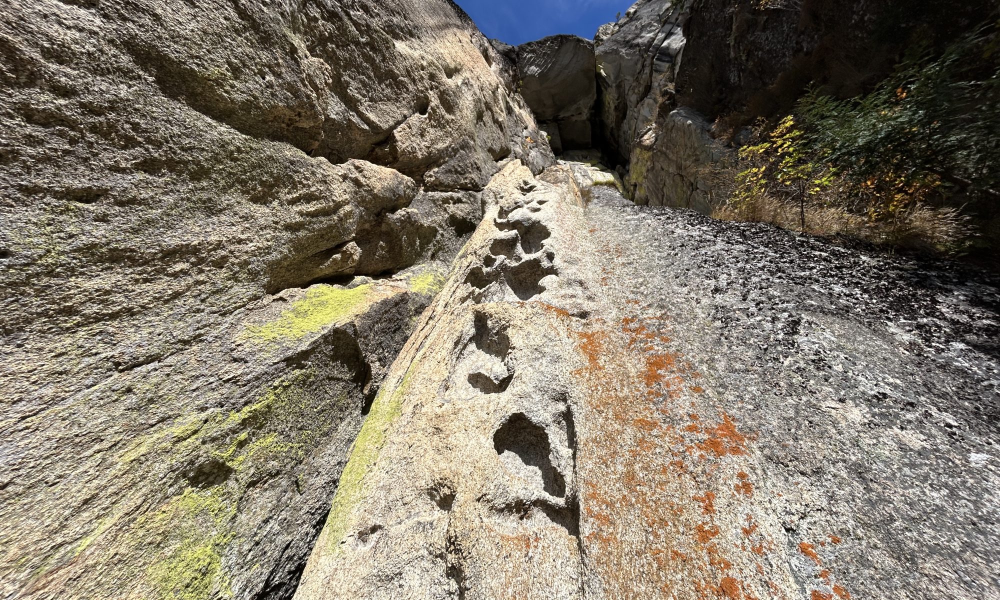 Narrow rocky canyon with high cliff walls, a sunlit groove in the center and patches of green lichen under a blue skyEXTREME-width trail through jagged granite canyon with lichens and a clear sky