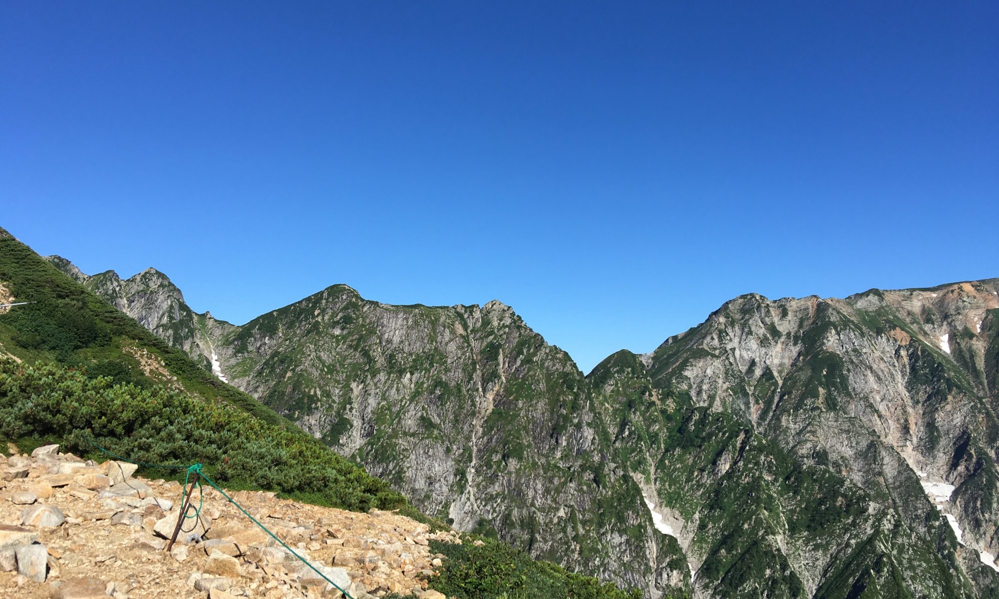 Rugged mountain ridge with rocky path in foreground, green shrubs, and a clear blue sky above