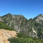 Rugged mountain ridge with rocky path in foreground, green shrubs, and a clear blue sky above