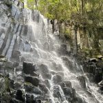 Waterfall cascading down a columnar basalt cliff with lush green vegetation at the top and edges, mist rising from the rocks.