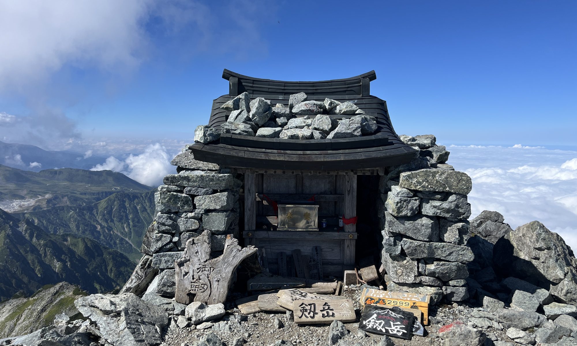Stone mountain hut atop a rocky peak with a wooden roof, surrounded by piled rocks and signboards at the front. (informative)