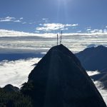 Peak of a mountain with two antennas at the summit, above a sea of clouds under a bright sun in a clear blue sky.