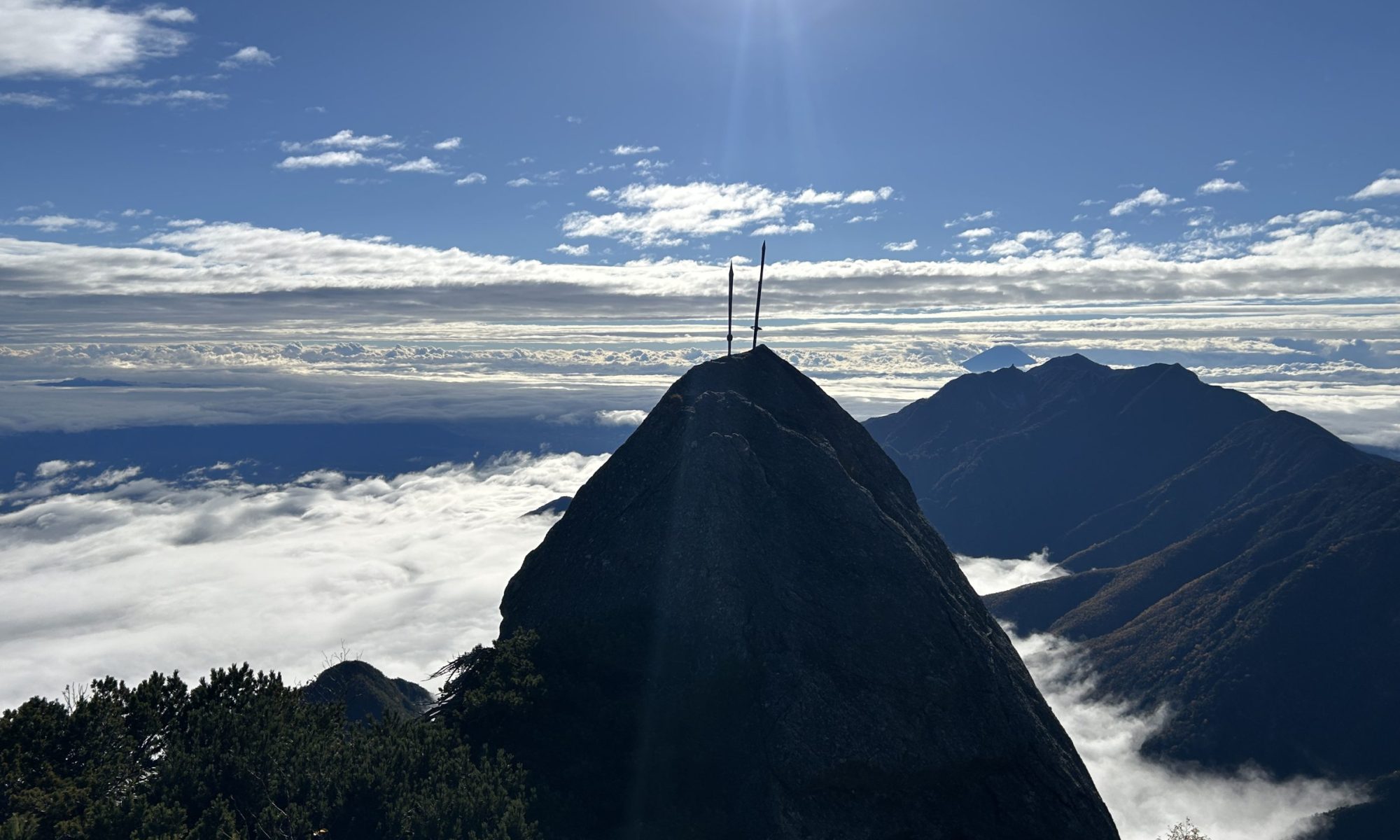 Peak of a mountain with two antennas at the summit, above a sea of clouds under a bright sun in a clear blue sky.