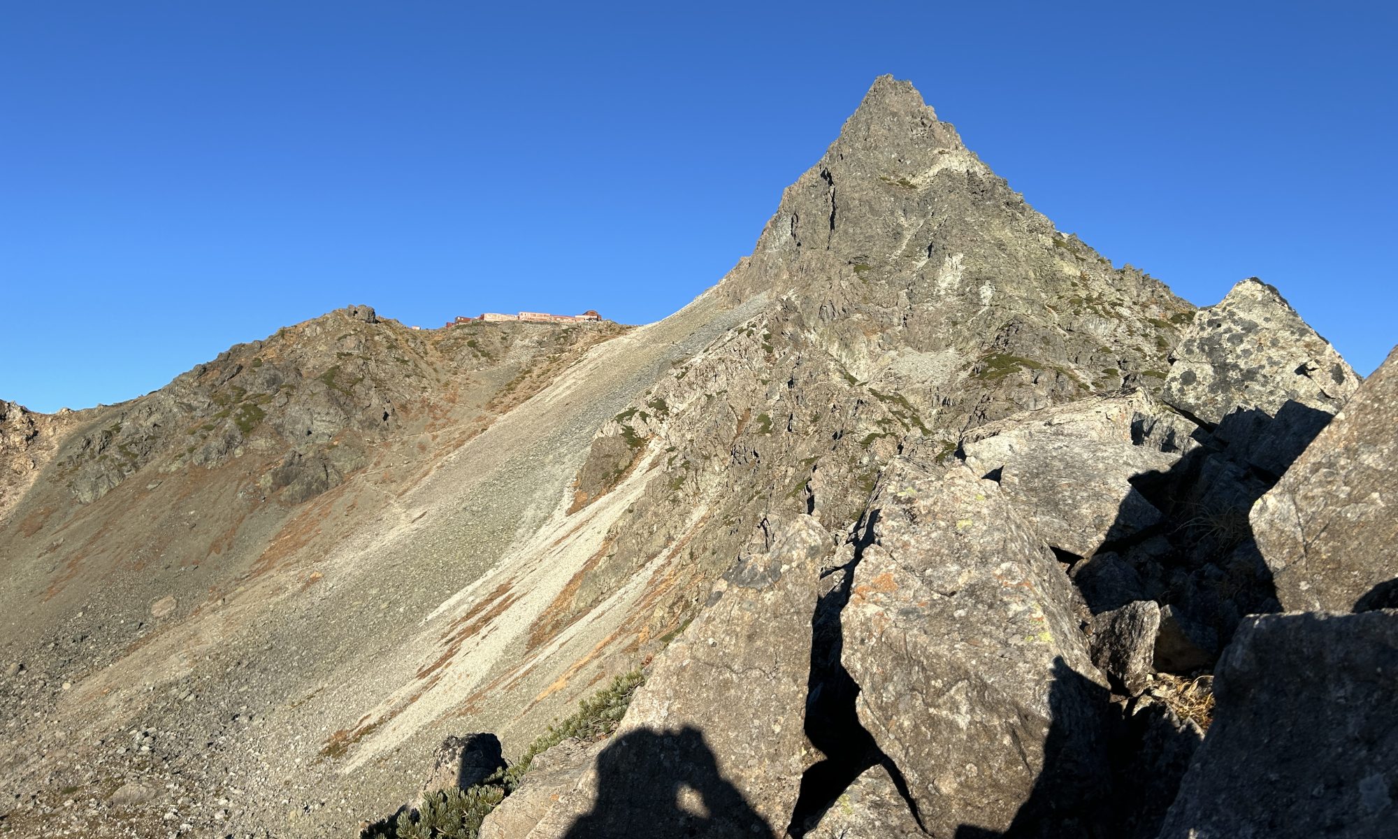 Rugged mountain ridge with a sharp peak under a clear blue sky; a small building sits on the left ridge.