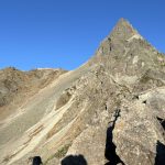 Rugged mountain ridge with a sharp peak under a clear blue sky; a small building sits on the left ridge.