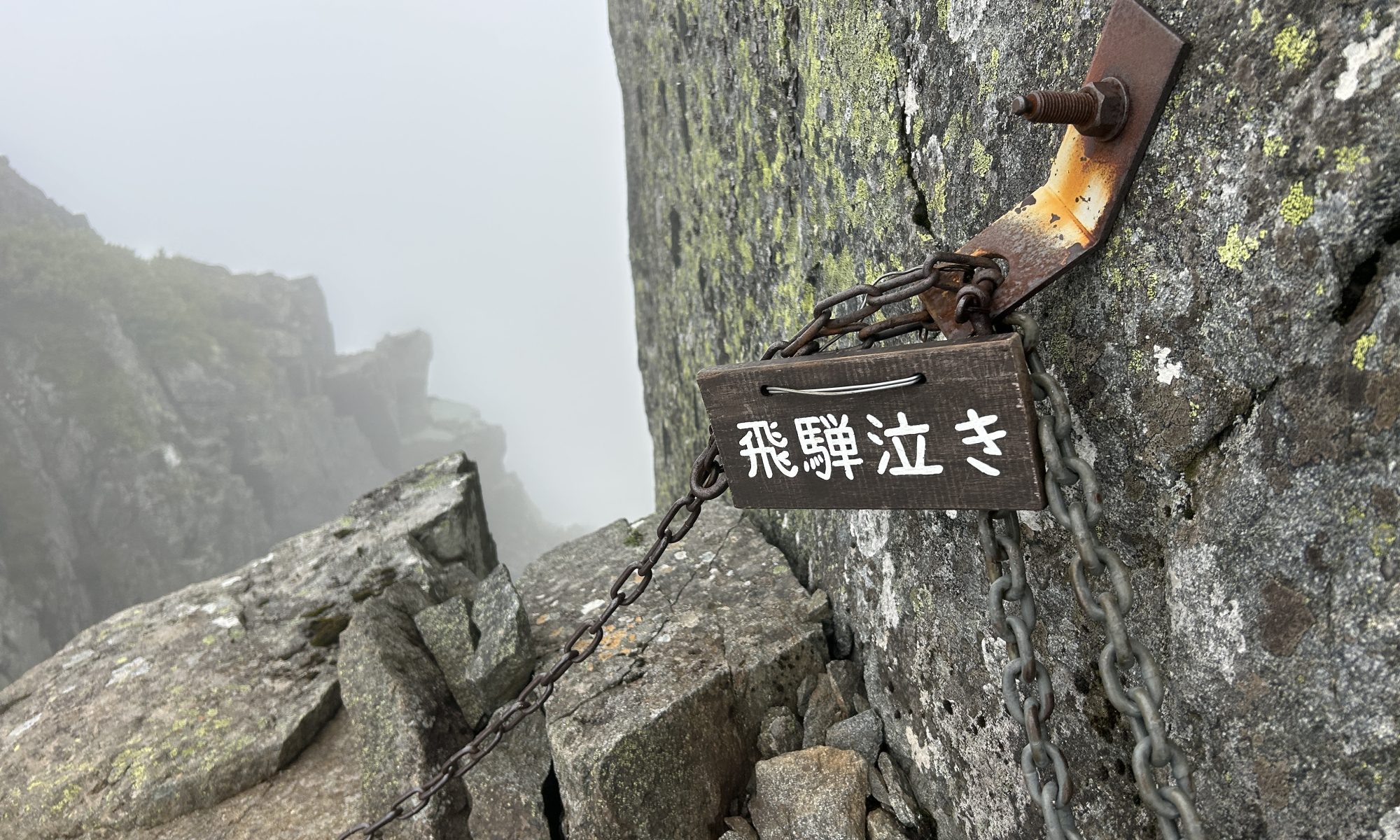 Wooden sign with Japanese characters 飛騨泣き attached to a chain across a sheer rock face on a misty mountain trail.
