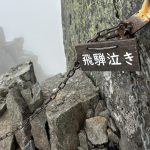 Wooden sign with Japanese characters 飛騨泣き attached to a chain across a sheer rock face on a misty mountain trail.